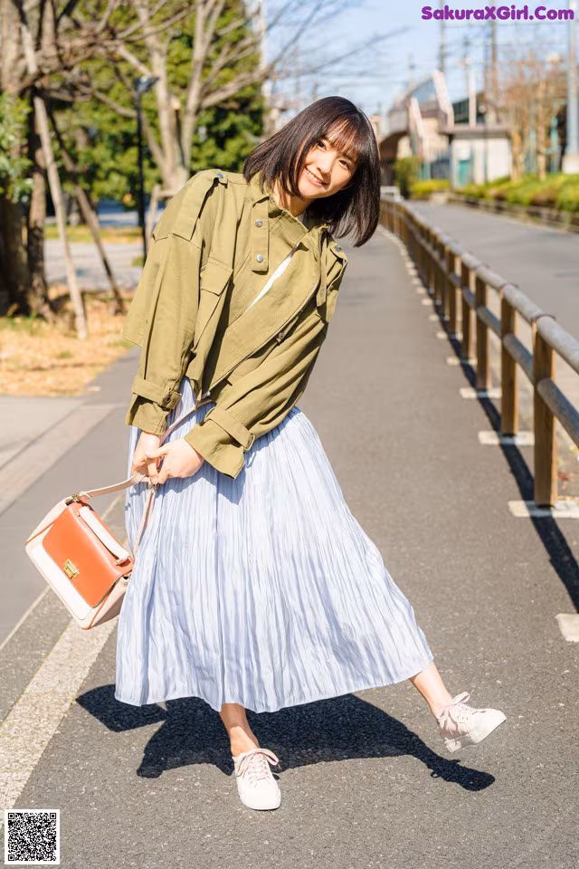 A woman in a green jacket and blue pleated skirt is walking down the street.