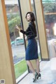A woman in a black shirt and blue skirt standing by a window.