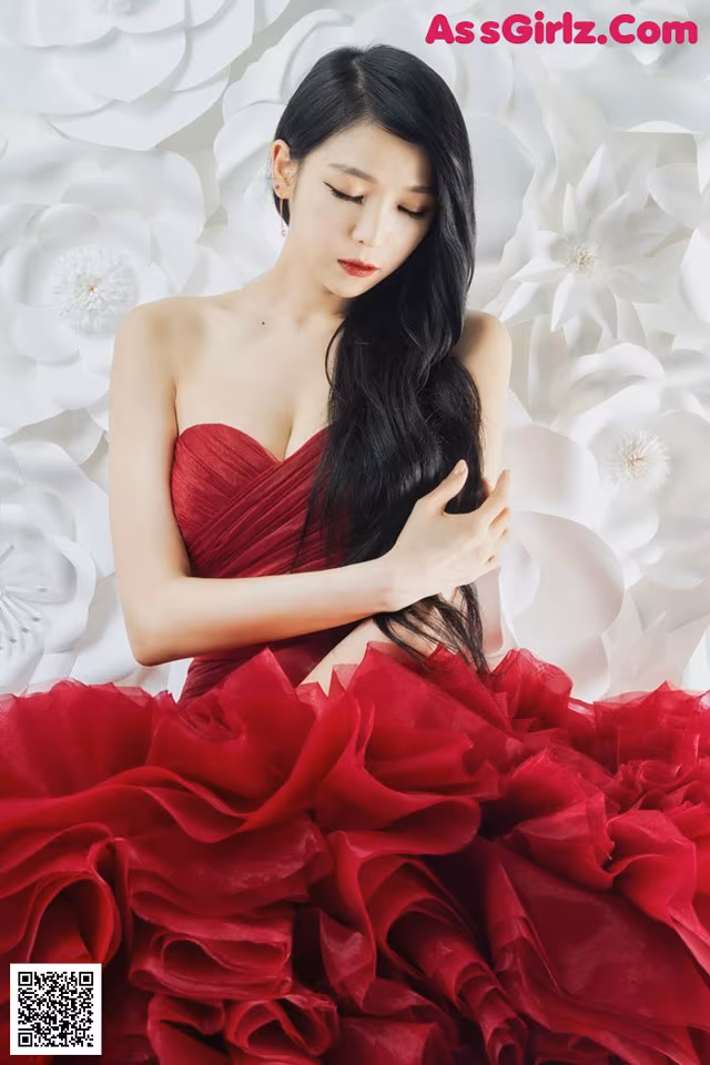 A woman in a red dress sitting in front of a wall of paper flowers.